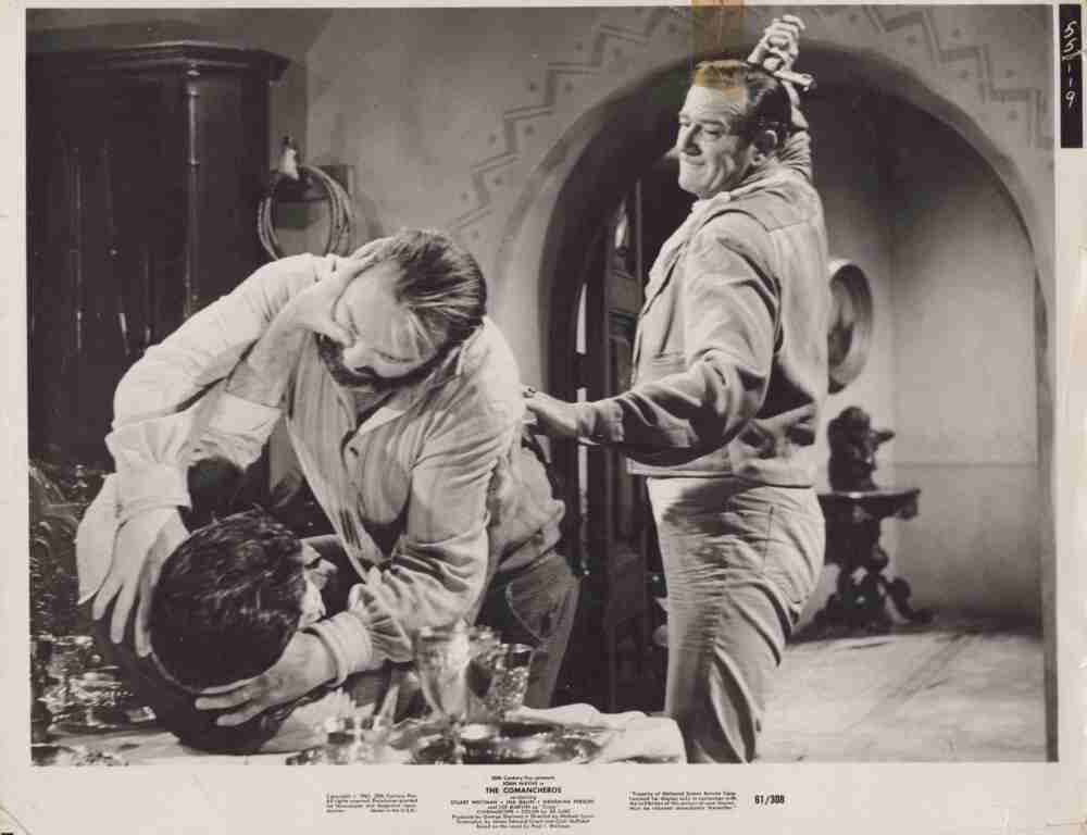 Two men wrestle on a table in a black-and-white studio scene while a third man watches, a prop sword on his head,”