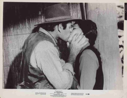 Man wearing a cowboy hat gently holds the woman's face as they passionately kiss beside a wooden wall.