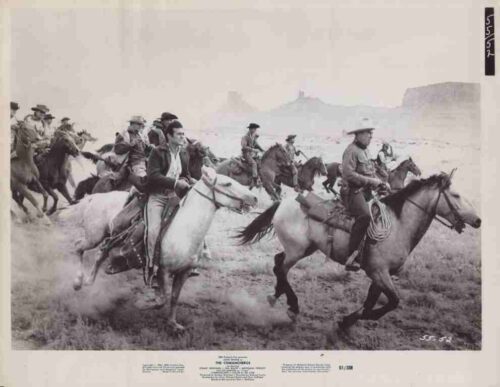 Cavalry soldiers riding horses across a dusty desert plain with mesas in the background.