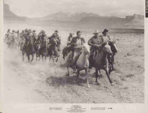 Group of cowboys on horseback riding in a dusty desert valley with mesas in the distance.