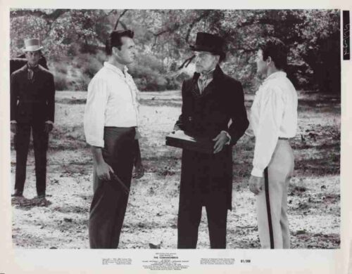 Three men in 19th‑century attire stand outdoors, one holding a book or clipboard while they converse.