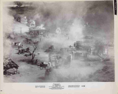 Historic black-and-white photo of a smoky outdoor camp with wagons, horses, and people tending fires near tents and structures.