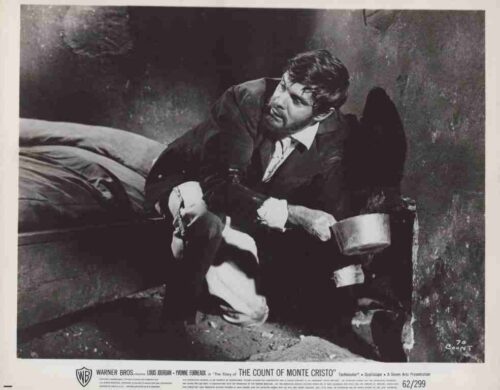 Man in 19th‑century clothing crouches beside a bed, holding a metal bowl, in a dimly lit scene from The Count of Monte Cristo.