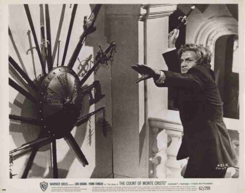 Man in a dark coat lunges forward, reaching toward a large spiked wheel mechanism mounted on the wall in a museum-like room.
