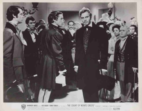 Movie still from The Count of Monte Cristo: a tall man in a dark suit stands among guests in 19th‑century attire in a crowded room.