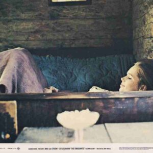 Woman resting in a wooden bed with a blue quilt, looking to the right; a white bowl sits on a table in the foreground.