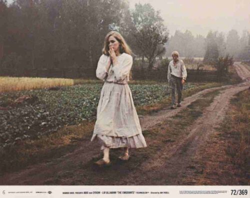 A woman in a white Victorian-style dress walks along a dirt rural road while a man follows behind in a field.