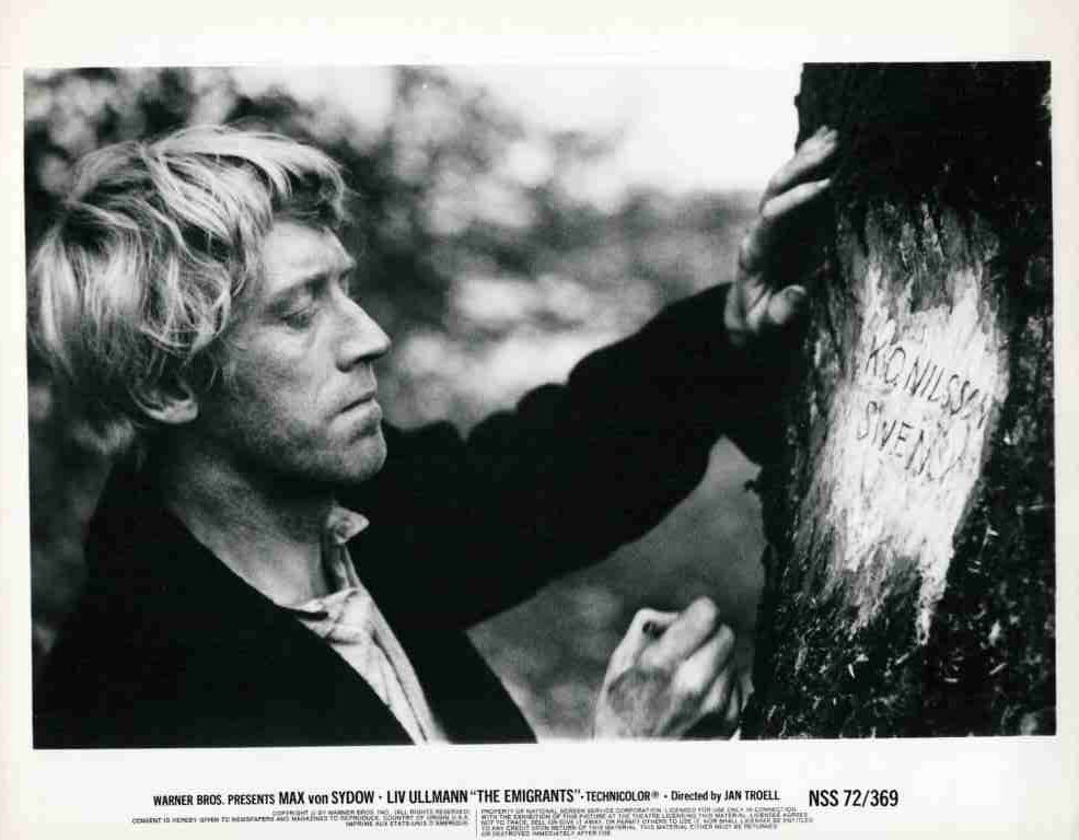 Black-and-white still of Max von Sydow as a man with light hair, touching a tree trunk and carving/writing on it outdoors in a somber scene from The Emigrants