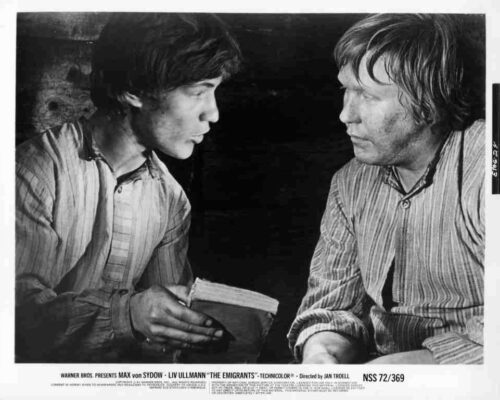 Two men in striped shirts sit facing each other, one holding a small book or notebook while talking.