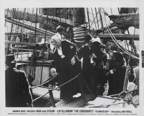 Black-and-white still from the film The Emigrants showing passengers on a ship deck with rigging; a woman helps a child down a gangplank as others wait nearby.