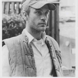 Black-and-white film still of a man wearing a cap and a padded vest, looking off to the side, in a wartime setting.