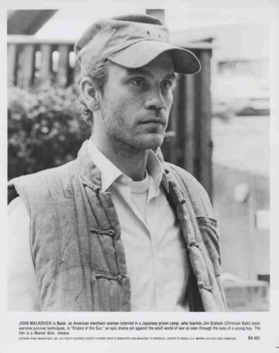 Black-and-white film still of a man wearing a cap and a padded vest, looking off to the side, in a wartime setting.