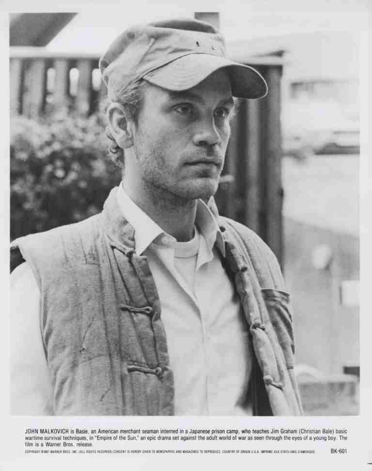 Black-and-white film still of a man wearing a cap and a padded vest, looking off to the side, in a wartime setting.