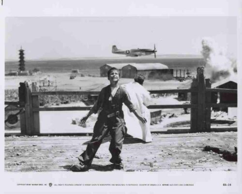 Boy in military uniform runs along a wooden seawall, with a beach and wartime structures in the background as a propeller plane flies overhead and smoke rises on the shore.