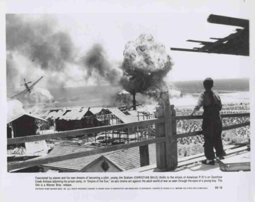 Boy on a seaside railing watches a beachfront air-attack scene with smoke and a burning plane in the distance.