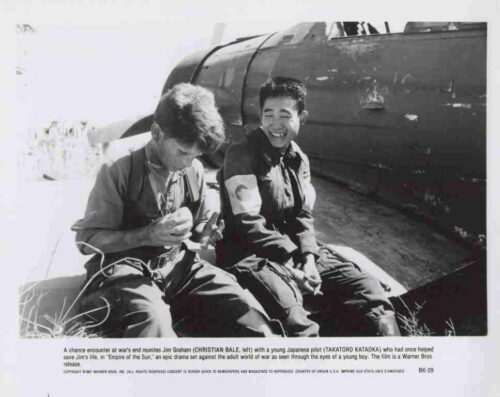 Two men in flight suits sit on the wing of a damaged aircraft, smiling at the camera in a desert airfield setting.