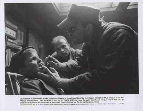 Black-and-white still from a war-era film: a boy sits with mouth open as a man in a cap and gloves examines him, another man watches from the background in a dim room.