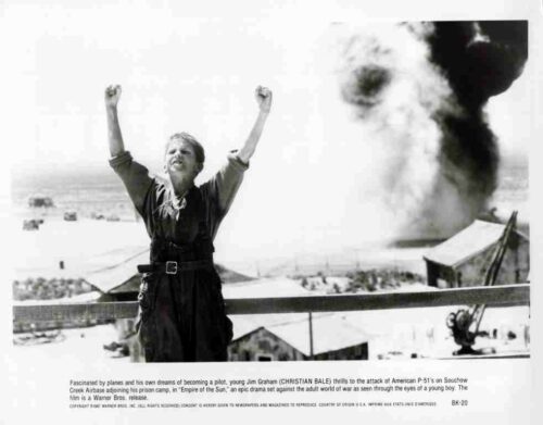 Young boy with arms raised in a desert camp as a large explosion rises behind him.