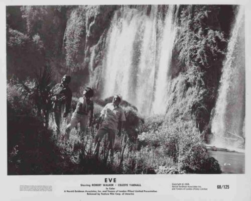 Three people stand among plants as a large waterfall cascades in the background.