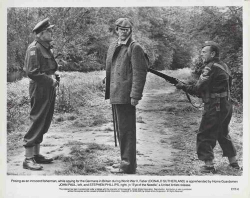 Movie still from Eye of the Needle: a man in a coat is held at gunpoint by two Home Guardsmen during WWII.