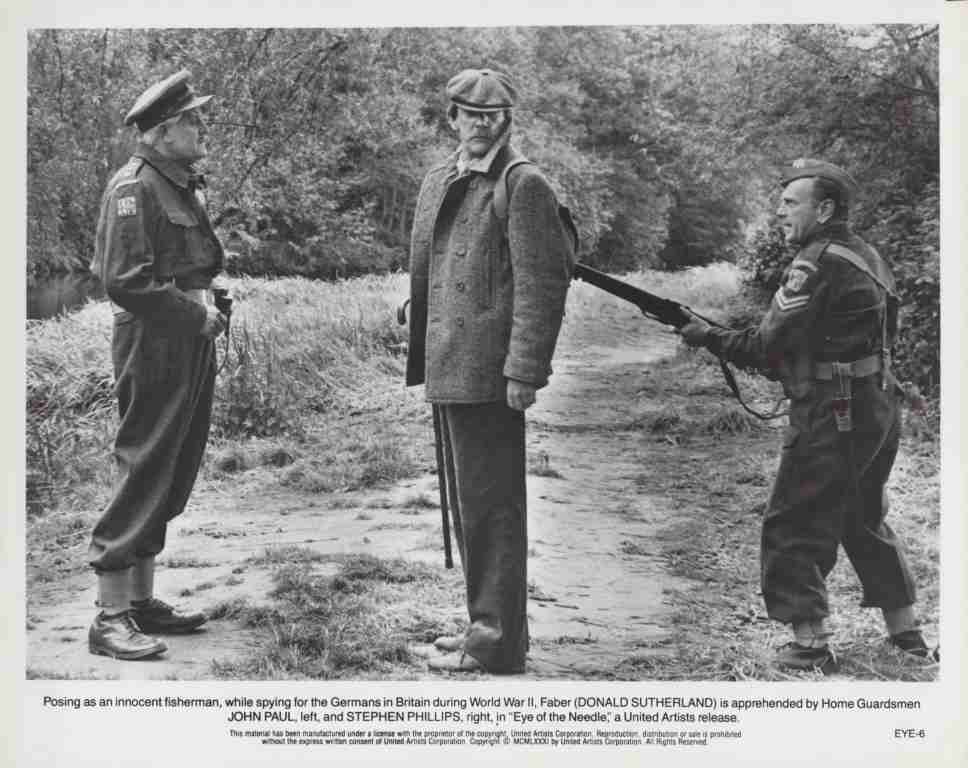 Movie still from Eye of the Needle: a man in a coat is held at gunpoint by two Home Guardsmen during WWII.