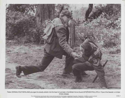 Man with a backpack kneels in a grassy area to hand a small object to a boy in uniform, during an outdoor drill in a wooded setting.