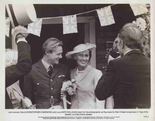 Bride and groom in vintage wedding outfits smile as they exit a doorway under Union Jack bunting, flanked by guests and a photographer.