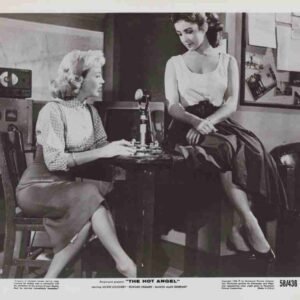 Two women in a vintage recording studio sit at a desk with a microphone between them and retro equipment in the background.