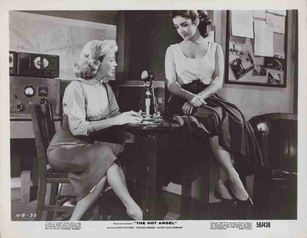 Two women in a vintage recording studio sit at a desk with a microphone between them and retro equipment in the background.