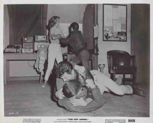 Two young people wrestle on the floor while others stand by near a desk with vintage radio equipment in a cluttered room.