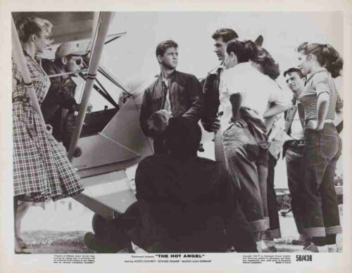 Movie lobby card for 'The Hot Angel' showing a group of people gathered around a small airplane, centering on a man in a leather jacket as others listen or converse nearby.