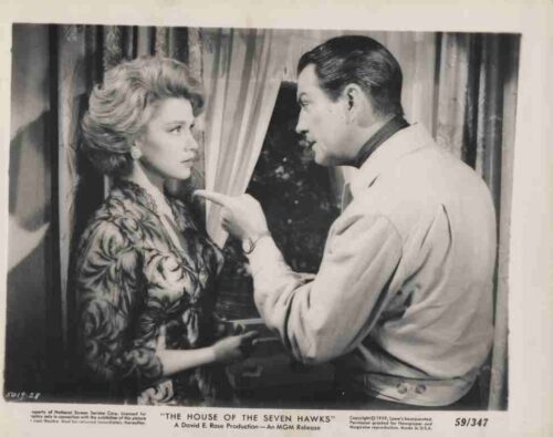 Still from a film showing a man in a light jacket pointing a finger at a woman's chin as they face each other by a window.