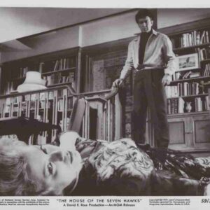 Man stands over a reclining woman on seating in a library filled with shelves and books, dramatic noir scene.