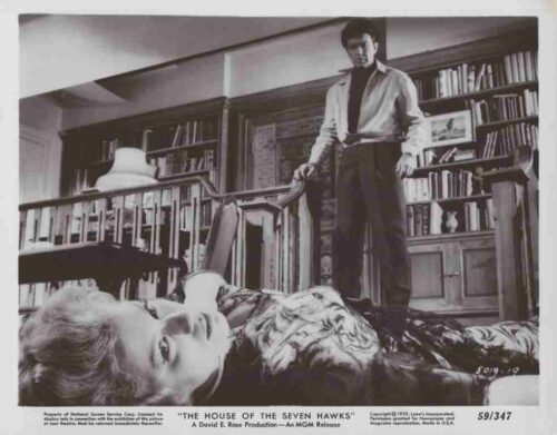 Man stands over a reclining woman on seating in a library filled with shelves and books, dramatic noir scene.