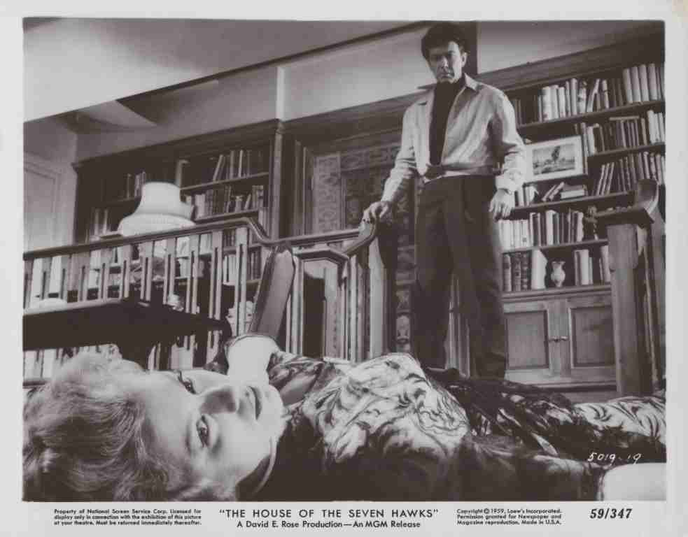 Man stands over a reclining woman on seating in a library filled with shelves and books, dramatic noir scene.