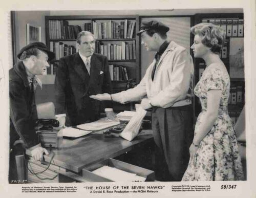 Black-and-white still from The House of the Seven Hawks: four people gathered around a desk in an office, reviewing papers as one man hands over a stack to another while a woman watches.