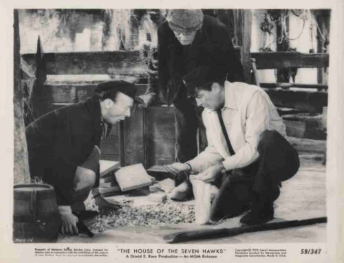 Black-and-white still from The House of the Seven Hawks: two men kneel and inspect a pile of rocks in a wooden outdoor setting, while a third man stands nearby.
