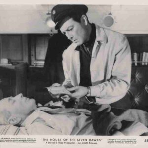 Black-and-white film still: a doctor in a cap and white coat leans over a bed, showing papers to an elderly man lying down (scene from The House of the Seven Hawks).