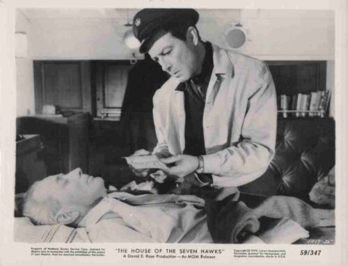 Black-and-white film still: a doctor in a cap and white coat leans over a bed, showing papers to an elderly man lying down (scene from The House of the Seven Hawks).