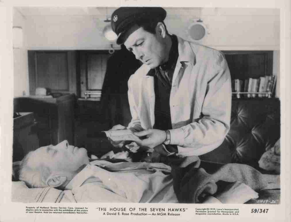 Black-and-white film still: a doctor in a cap and white coat leans over a bed, showing papers to an elderly man lying down (scene from The House of the Seven Hawks).