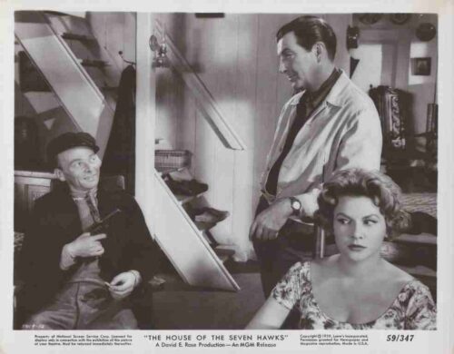 Movie still from The House of the Seven Hawks: a man in a cap sits by a staircase, another man stands nearby, and a woman with curled hair sits in the foreground.