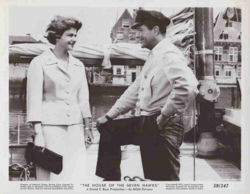 Two people on a dock: a smiling woman in a light suit stands while a man in a cap rests one knee on a railing beside a sailboat, mid-conversation.