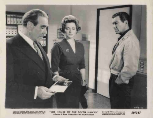 Three adults stand in a mid-century room: a suited man shows a photo to a woman, while a man in a light shirt leans against a doorway, all looking serious.