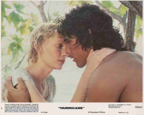 Two topless adults lean in close, foreheads touching, sharing an intimate moment under a tree on a sunny beach.