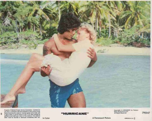 Man carrying a woman in a white dress along a tropical beach with palm trees in the background.