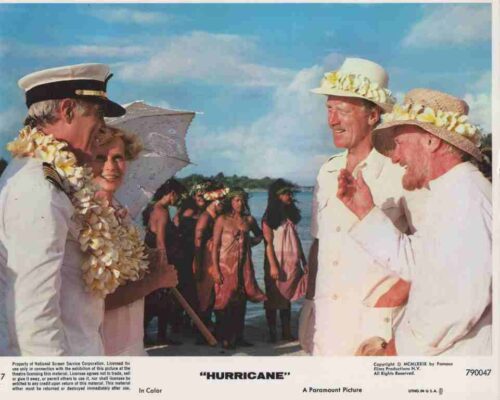 Three men in white outfits with flower leis greet each other on a sunny beach, extras in colorful dress in the background.