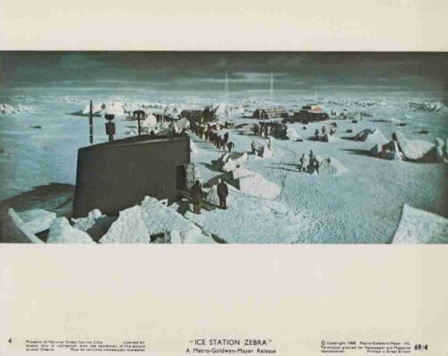 Film still of an Antarctic ice station: a dark rectangular shelter amid tents and crew on a snowy, wind-swept landscape.