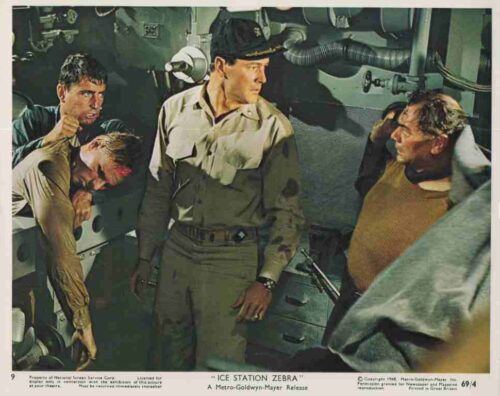 Three sailors in a submarine’s control room, with tense expressions and gear surrounding them, centered on a man in a light uniform looking ahead.