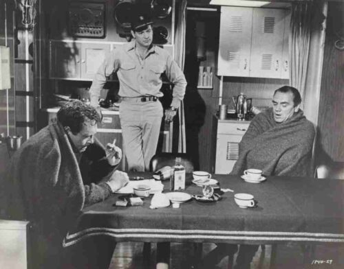 Three men are gathered around a table in a small kitchen; two are wrapped in blankets, with cups and a bottle on the table, while a uniformed man stands nearby.