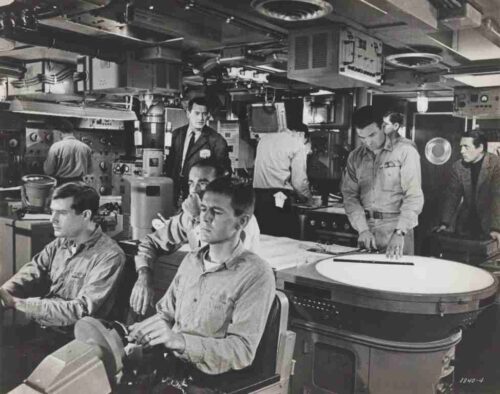 Historic black-and-white photo of engineers in a crowded 1960s control room, working at consoles and gauges alongside large machinery.
